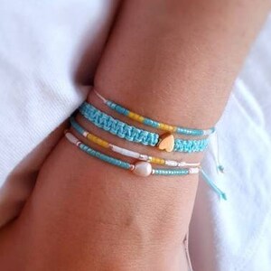 Può includere: Un set di quattro braccialetti al polso di una persona. I braccialetti sono fatti di filo blu, bianco e giallo. Un braccialetto ha un piccolo ciondolo a forma di cuore dorato.
