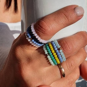 May include: A stack of colourful beaded rings on a finger. The rings are made of small, round beads in shades of blue, green, yellow, and white. The rings are worn on the index finger of a hand holding a white mug.