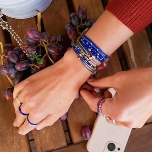 Puede incluir: Primer plano de la muñeca de una persona que lleva una pila de pulseras de cuentas azules y doradas. Las pulseras están superpuestas y tienen pequeños abalorios. La mano de la persona está apoyada sobre una superficie de madera con uvas en el fondo.