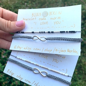 Puede incluir: Dos pulseras de amistad grises con dijes de infinito plateados. Las pulseras están hechas con pequeñas cuentas y tienen el mensaje "I LOVE YOU" en código morse en las tarjetas.
