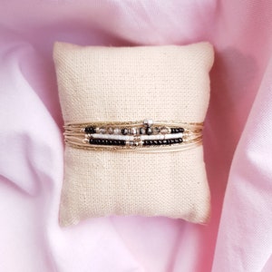 Op de afbeelding: Een stapel gouden, zwarte en witte kralen armbanden op een beige stoffen displaystandaard.