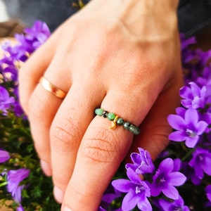 Może przedstawiać: Złoty pierścionek z małym wisiorkiem i zielonymi koralikami z kamieni szlachetnych. Pierścionek jest na palcu, a dłoń spoczywa na fioletowych kwiatach.