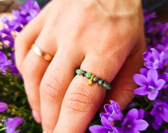Könnte beinhalten: Ein goldener Ring mit einem kleinen Anhänger und grünen Edelsteinperlen. Der Ring befindet sich an einem Finger und die Hand ruht auf violetten Blumen.