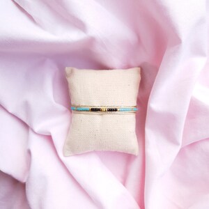 May include: Two beaded bracelets on a white pillow. The top bracelet is black and gold with turquoise beads. The bottom bracelet is gold with turquoise beads.