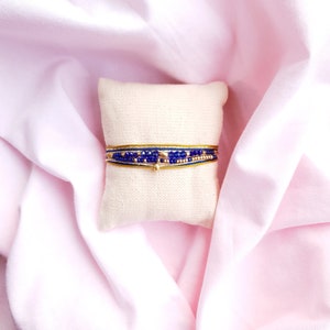 May include: A stack of three bracelets with blue and gold beads on a white cushion. The bracelets are made of different materials, including beads, string, and metal.