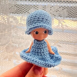 May include: A small doll wearing a light blue crocheted hat and dress. The doll has large eyes and a smiling expression. The crochet work appears handmade, with visible stitch patterns. The doll is held in a hand.