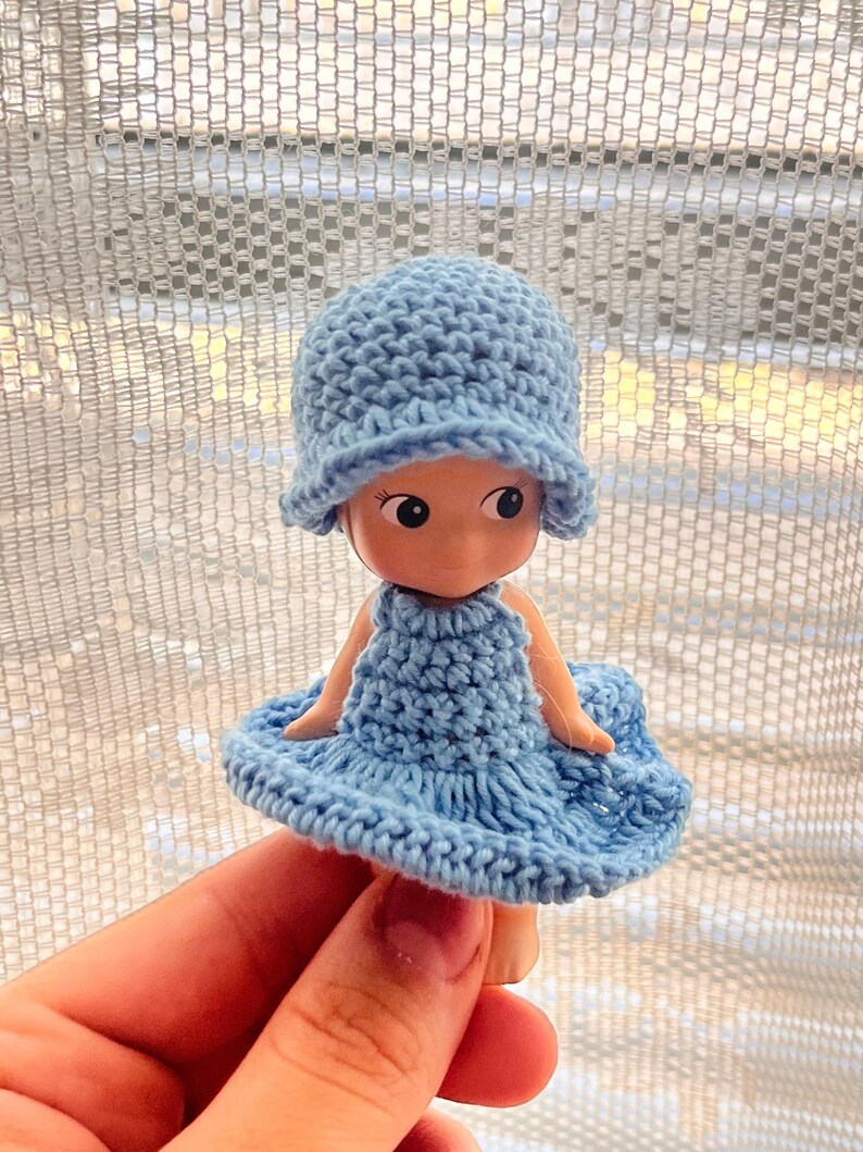 May include: A small doll wearing a light blue crocheted hat and dress. The doll has large eyes and a smiling expression. The crochet work appears handmade, with visible stitch patterns. The doll is held in a hand.