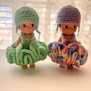 May include: Two small dolls wearing handmade crochet outfits. One doll is dressed in a mint green outfit, including a hat and a ruffled skirt. The other doll is wearing a multi-colored outfit with pink, blue, and yellow hues. Both dolls have a similar facial design.