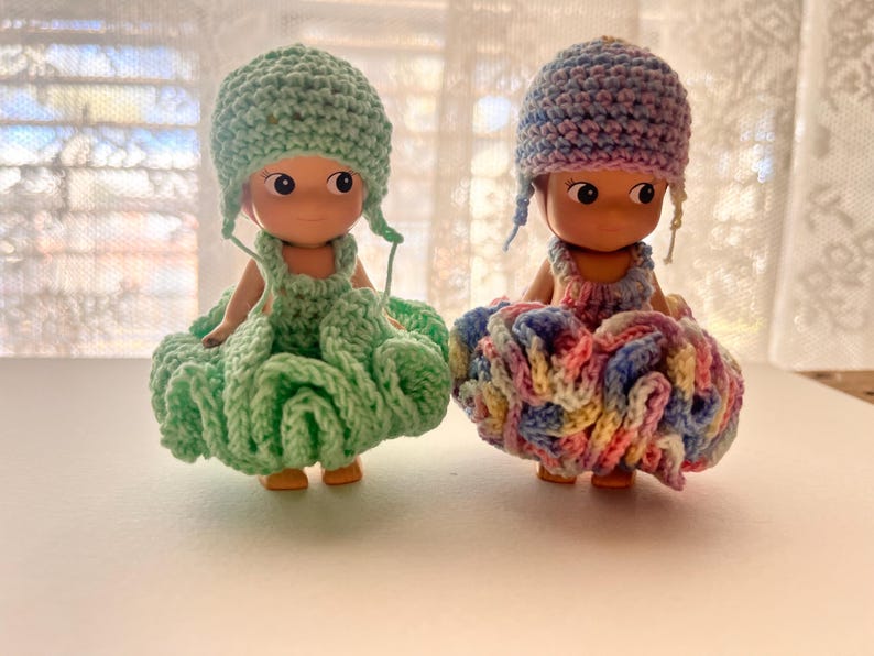 May include: Two small dolls wearing handmade crochet outfits. One doll is dressed in a mint green outfit, including a hat and a ruffled skirt. The other doll is wearing a multi-colored outfit with pink, blue, and yellow hues. Both dolls have a similar facial design.