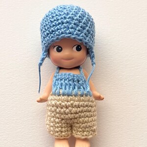May include: A small doll wearing a light blue crocheted hat with ear flaps, a light blue and white crocheted top, and tan crocheted shorts. The doll has a smiling expression and is set against a white background.