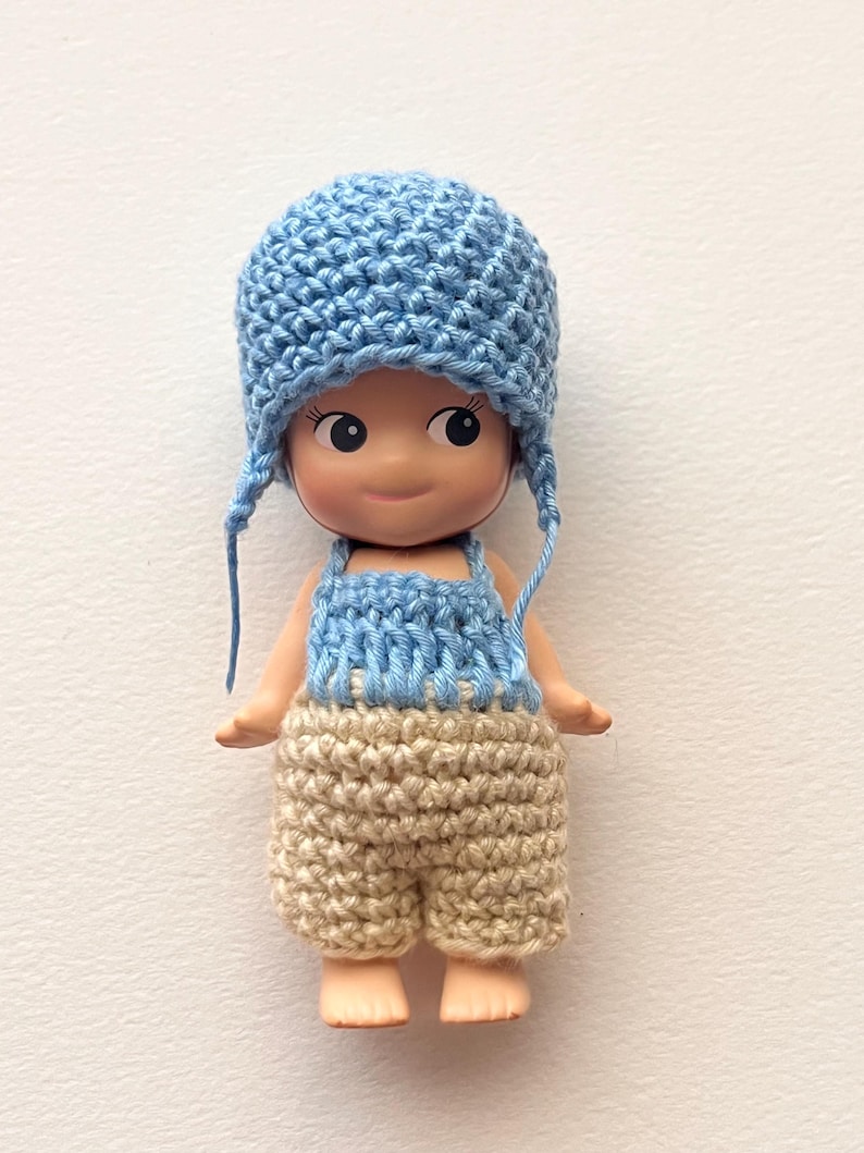 May include: A small doll wearing a light blue crocheted hat with ear flaps, a light blue and white crocheted top, and tan crocheted shorts. The doll has a smiling expression and is set against a white background.