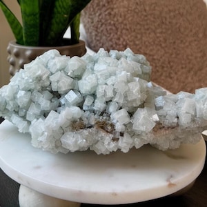 May include: A large, pale blue crystal cluster with a cubic structure, displayed on a white marble pedestal. The crystal has a rough, textured surface and is surrounded by a neutral background. The overall theme is natural decor.