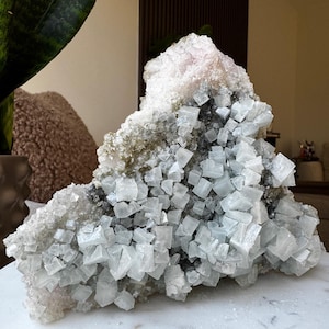 May include: A large, natural crystal formation with a complex structure. The crystal features a mix of white, gray, and pale pink hues. The surface is composed of numerous small, cube-shaped crystals, creating a textured appearance. The crystal sits on a white marble surface.