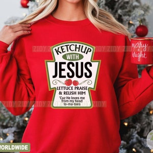 May include: A red sweatshirt with a ketchup bottle graphic that reads "Ketchup with Jesus." The graphic includes the phrases "Lettuce Praise & Relish Him" and "'Cuz He loves me from my head to-ma-toes." A red mug with "merry + bright" is held in the hand.
