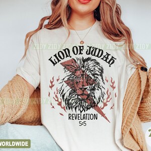 以下が含まれることがあります： クリーム色のTシャツ。 荒れたライオンの頭のグラフィック、稲妻、炎が特徴です。「LION OF JUDAH」と「REVELATION 5:5」の文字がプリントされています。 デザインは黒と赤です。 衣料品です。