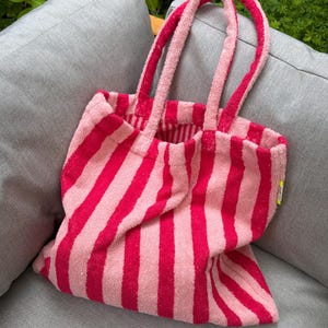 Könnte beinhalten: Eine Tragetasche aus Frottee mit vertikalen Streifen in abwechselnden Farbtönen von Pink und Hellrosa. Die Tasche verfügt über zwei passende Stoffgriffe und ein kleines gelbes Etikett, das an der Seitennaht des Hauptkörpers befestigt ist.