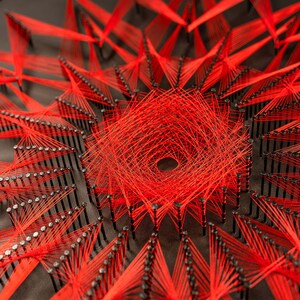 May include: A close-up of a red string art piece with a circular design. The string is wrapped around nails hammered into a dark surface, creating a geometric pattern.