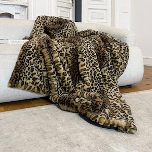 Może przedstawiać: Brązowo-beżowy koc w panterkę, drapowany na białej sofie. Koc ma miękką, puszystą fakturę i jest wykonany z imitacji futra.