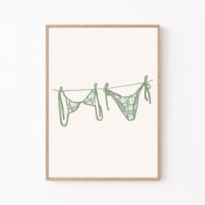 May include: A framed print of a green floral patterned bikini hanging on a clothesline. The print is on a white background.