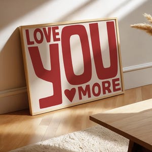 May include: A framed print with the text "LOVE YOU MORE" in red. The artwork includes a heart symbol and is set against a cream background. The frame is light wood-toned. The print is leaning against a wall, suggesting a home decor item.