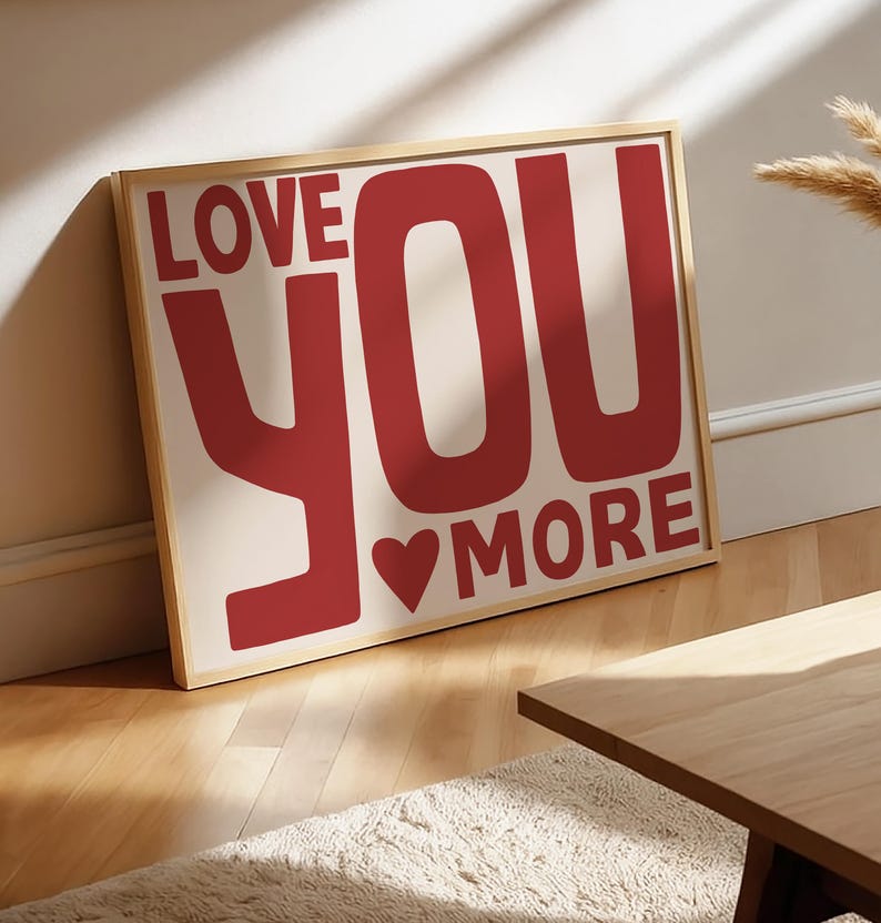 May include: A framed print with the text "LOVE YOU MORE" in red. The artwork includes a heart symbol and is set against a cream background. The frame is light wood-toned. The print is leaning against a wall, suggesting a home decor item.
