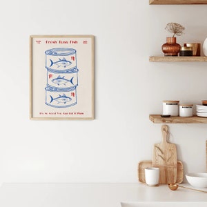 May include: A framed print featuring three stacked tuna cans with blue illustrations of fish. The text "Fresh Tuna Fish" and "It's So Good You Can Eat It Plain" are in red. The print has a beige background and a wooden frame.