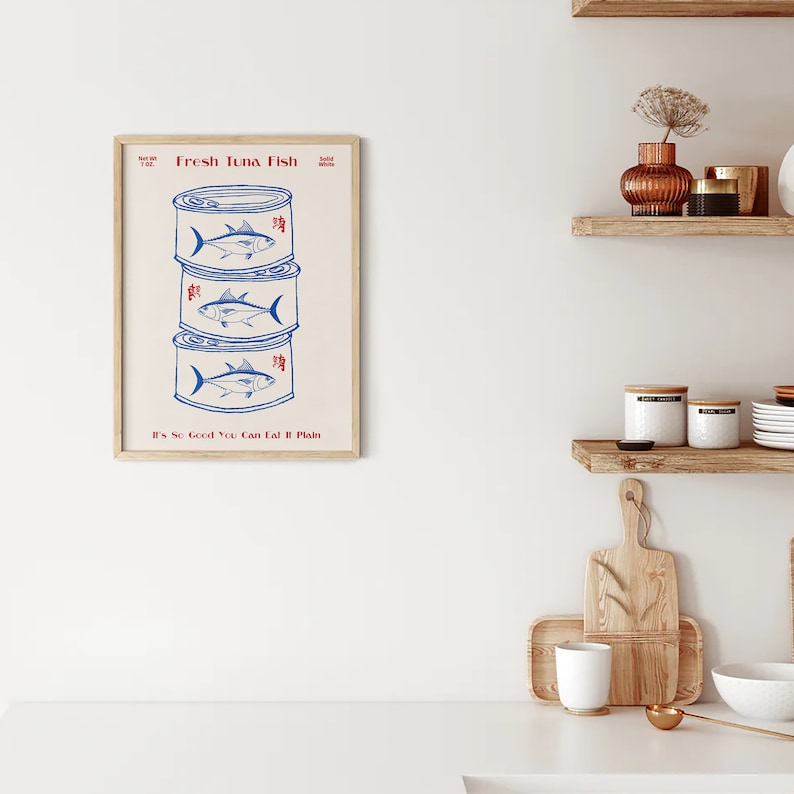 May include: A framed print featuring three stacked tuna cans with blue illustrations of fish. The text "Fresh Tuna Fish" and "It's So Good You Can Eat It Plain" are in red. The print has a beige background and a wooden frame.