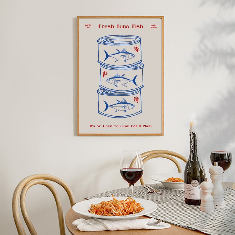 May include: A framed print featuring a stack of three blue and white tuna cans with a fish illustration. The text "Fresh Tuna Fish" and "It's So Good You Can Eat It Plain" are in red. A dining table with pasta, wine, and a candle is in the foreground.