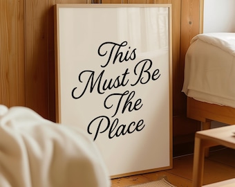 May include: A framed print featuring the phrase "This Must Be The Place" in black cursive lettering. The artwork is set against a light backdrop, framed in a natural wood tone, and positioned against a wooden wall.