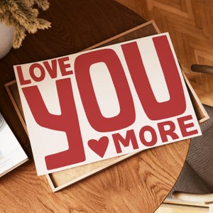 May include: A rectangular sign with the words "LOVE YOU MORE" in bold, red letters. A red heart is between "YOU" and "MORE". The sign is on a wooden surface.