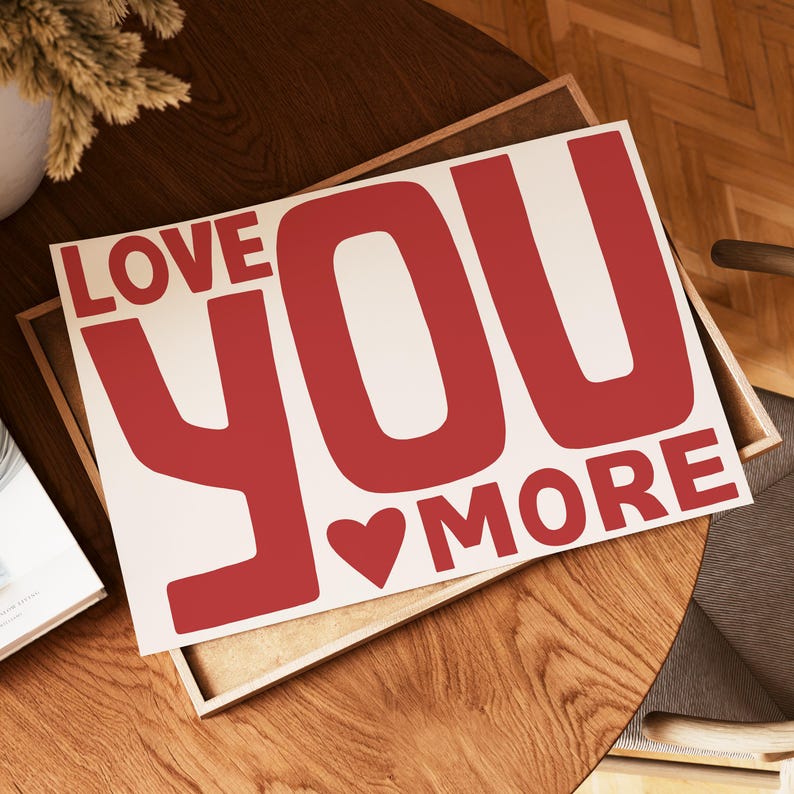 May include: A rectangular sign with the words "LOVE YOU MORE" in bold, red letters. A red heart is between "YOU" and "MORE". The sign is on a wooden surface.