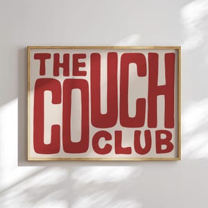 Può includere: Una stampa artistica incorniciata con le parole "THE COUCH CLUB" in lettere rosse e in grassetto. Il testo è centrato su uno sfondo color crema e la cornice è di colore legno chiaro. L'opera d'arte è esposta su una parete bianca.