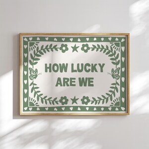May include: A framed print with the text "HOW LUCKY ARE WE" in green. The text is centered on a cream-colored background, surrounded by a green border with floral and butterfly motifs. The frame is a light brown color.