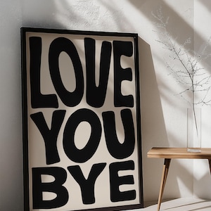 May include: Black framed art print with the words "LOVE YOU BYE" in bold, black, rounded letters on a cream background. The artwork is displayed against a white wall, with a wooden table and a vase with branches.