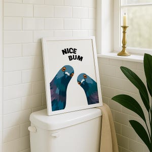 Op de afbeelding: Een ingelijste print met twee cartoonduiven met blauwe en paarse veren, oranje ogen en de tekst "NICE BUM" in het zwart. De print wordt weergegeven op een wit toilet.