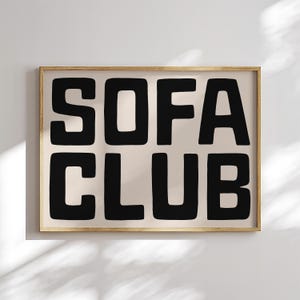 May include: A framed art print with the words "SOFA CLUB" in bold, black letters. The text is centered on a beige background, encased in a thin, light brown wooden frame. The artwork is a minimalist design.