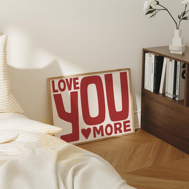 May include: A framed print with the text "LOVE YOU MORE" in red, with a heart symbol. The artwork is displayed on a light-colored wall, leaning against a bed and a wooden floor.
