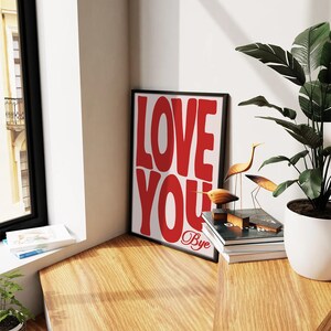 K&ouml;nnte beinhalten: Ein gerahmter Druck mit den Worten "LOVE YOU" in gro&szlig;en roten Buchstaben. Die Worte "Bye" sind darunter in Kursivschrift geschrieben. Der Druck h&auml;ngt an einer Wand neben einer Topfpflanze und einem Stapel B&uuml;chern.