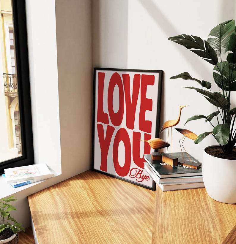 K&ouml;nnte beinhalten: Ein gerahmter Druck mit den Worten "LOVE YOU" in gro&szlig;en roten Buchstaben. Die Worte "Bye" sind darunter in Kursivschrift geschrieben. Der Druck h&auml;ngt an einer Wand neben einer Topfpflanze und einem Stapel B&uuml;chern.