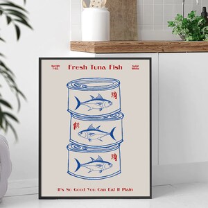 May include: A framed print featuring a stack of three blue and white tuna fish cans. The words "Fresh Tuna Fish" are above the cans, and "It's So Good You Can Eat It Plain" is below. The net weight is 7 oz.