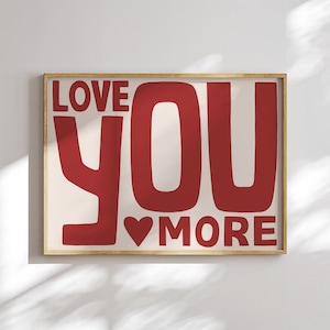 May include: A framed print featuring the phrase "LOVE YOU MORE" in large, red letters. The word "YOU" is central, with a red heart replacing the "O" in "MORE". The background is a pale cream colour.