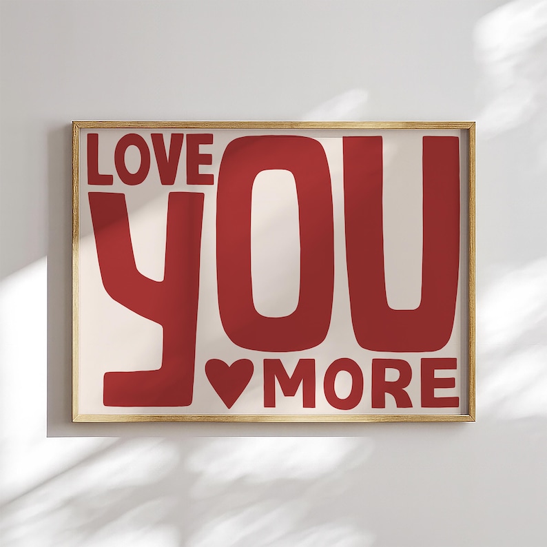 May include: A framed print featuring the phrase "LOVE YOU MORE" in large, red letters. The word "YOU" is central, with a red heart replacing the "O" in "MORE". The background is a pale cream colour.