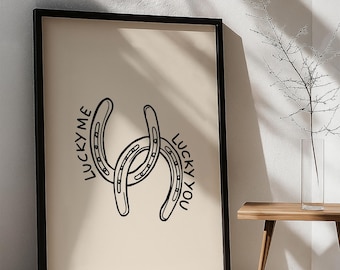 May include: Black framed art print featuring two intertwined horseshoe illustrations. The words "LUCKY ME" and "LUCKY YOU" are written on the print. The background is a light beige color.