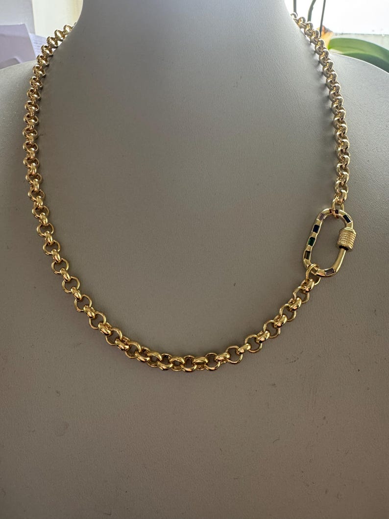 May include: A gold-colored chain necklace with a large, decorative clasp. The necklace is made of round, interlocking links. The clasp is a large oval shape with a screw closure.