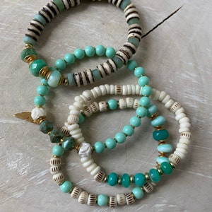May include: Set of four bracelets featuring white and green beads. The bracelets are crafted from a variety of materials, including wood, glass, and metal. The bracelets are layered on top of each other.