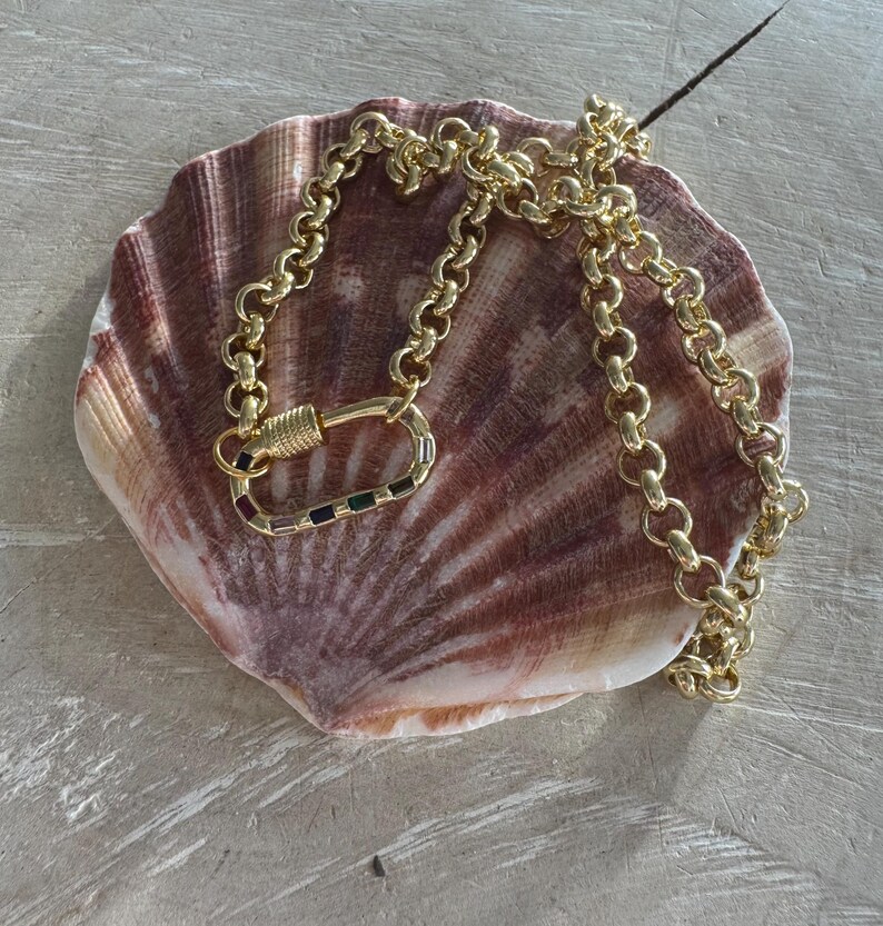 May include: A gold-colored chain necklace with a large clasp featuring colorful rectangular accents. The necklace is displayed on a large, textured seashell with shades of brown and pink.