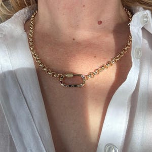 May include: Gold-toned chain necklace with a unique clasp. The necklace features a series of rounded links and a rectangular clasp with a decorative element. The jewelry is worn against a white shirt.