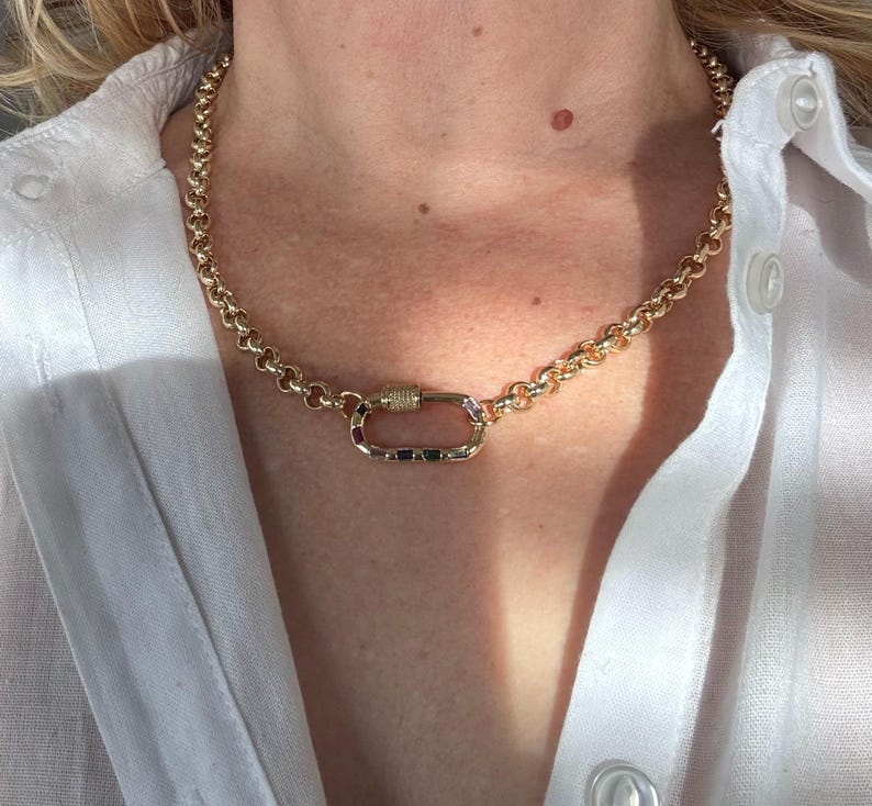 May include: Gold-toned chain necklace with a unique clasp. The necklace features a series of rounded links and a rectangular clasp with a decorative element. The jewelry is worn against a white shirt.