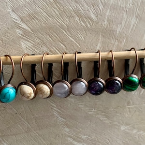 Puede incluir: Un conjunto de ocho pares de pendientes con piedras de diferentes colores. Los pendientes están hechos de cobre y tienen un diseño simple de aro. Las piedras son turquesa, blancas, morado claro, morado oscuro, verde y malaquita.