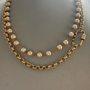 May include: A gold-toned necklace with two strands. The top strand features cream-colored pearls, while the bottom strand is a chunky chain. The necklace is fastened with a gold clasp with black and green accents.